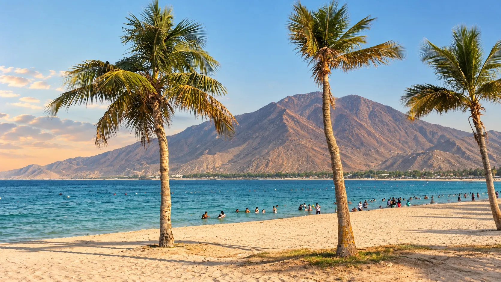 Ras Al Khaimah beach framed by palm trees, with blue water and the Hajar Mountains rising behind it.