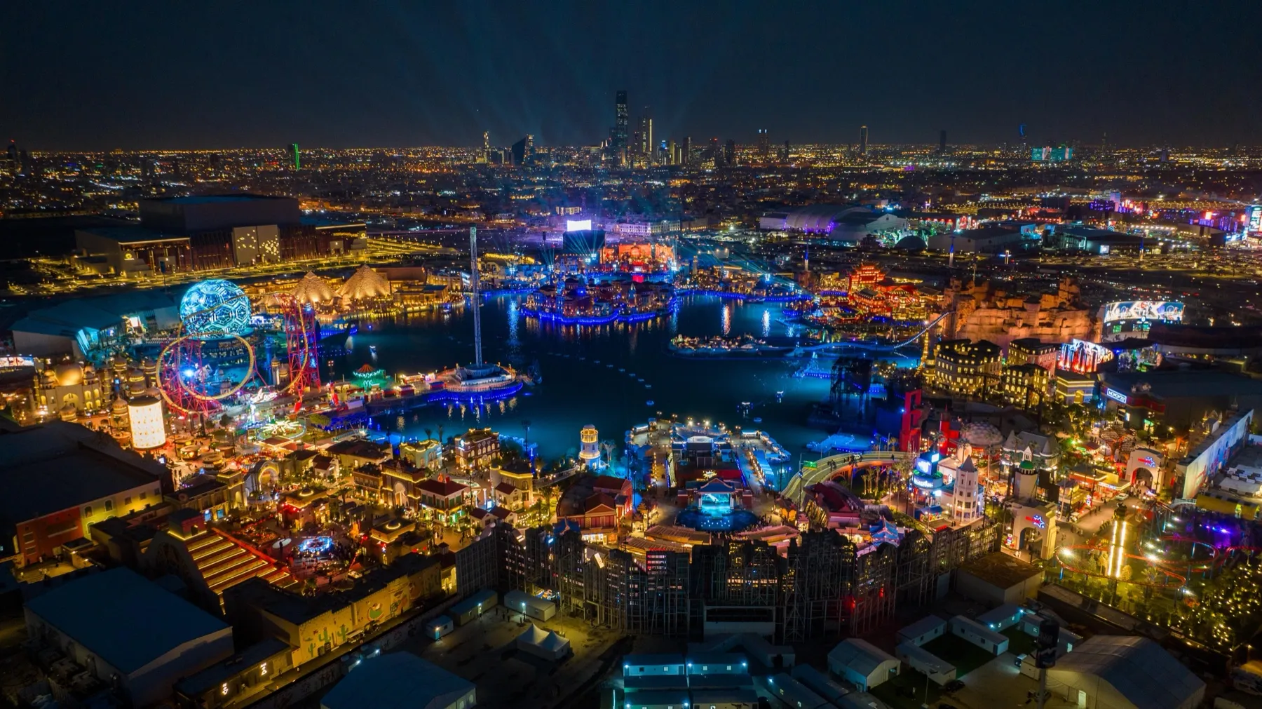 An aerial night view of Boulevard World in Riyadh, with its lake, illuminated pavilions and rides
