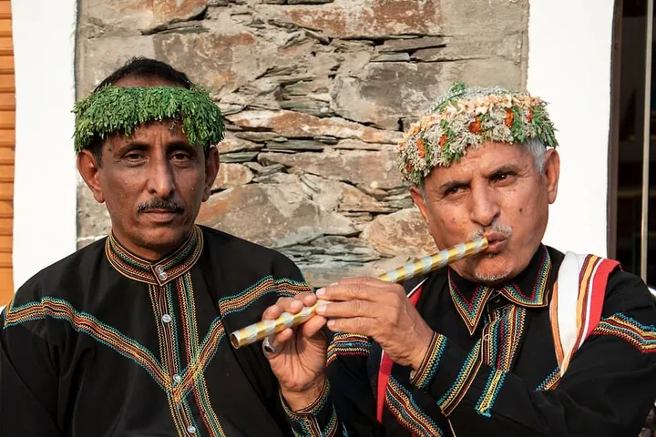 An Asir musician in traditional dress plays a flute beside another performer