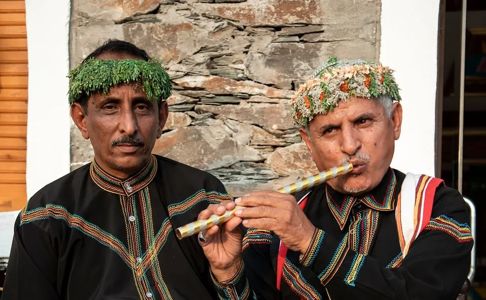 An Asir musician in traditional dress plays a flute beside another performer